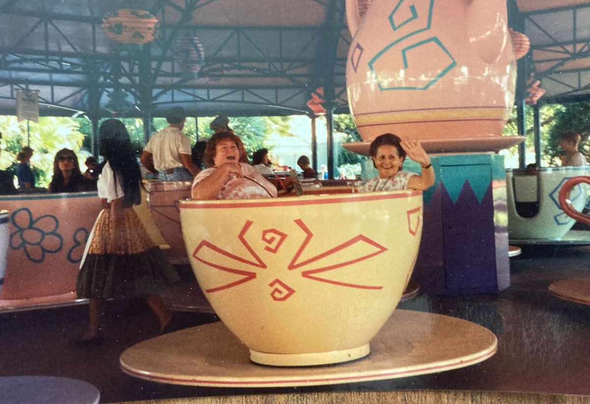 two adult women, mother & daughter, smiling and having fun on the tea cups ride in Disney World. They are centered in a yellow cup with orange decoration and other people are in different colored cups in the background