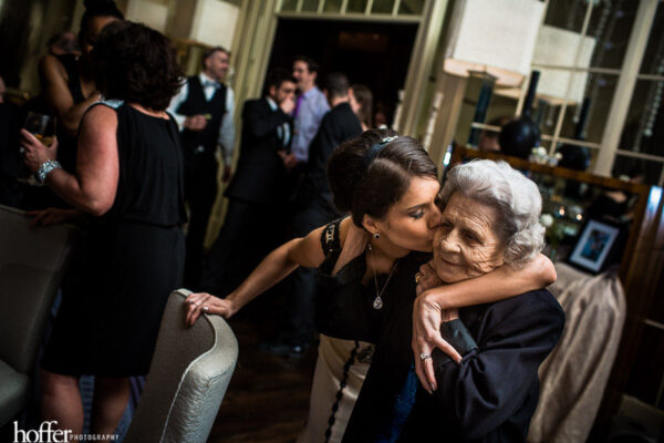 a bride kisses her shorter, older grandmother with a wedding party going on in the background
